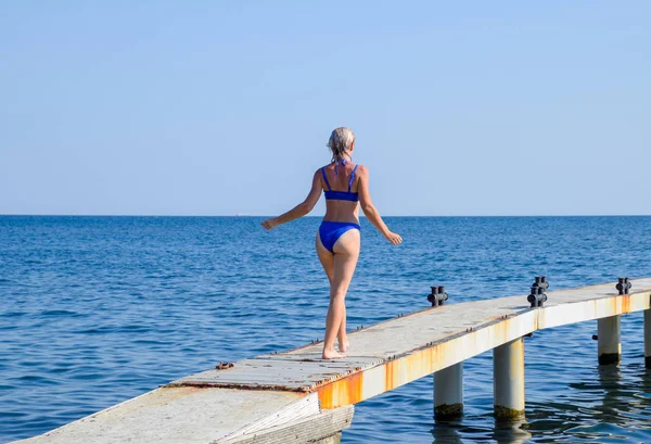 Красивая девушка в синем бикини идёт к морю. Бетонный причал. Прыжки в воду с пирса. Девчонки — стоковое фото