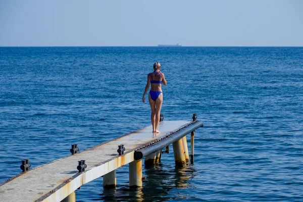 Ein schönes Mädchen im blauen Bikini geht die Seebrücke zum Meer entlang. Schiffsbeton-Pier. Sprung von der Seebrücke ins Wasser. schöne Beute Mädchen — Stockfoto