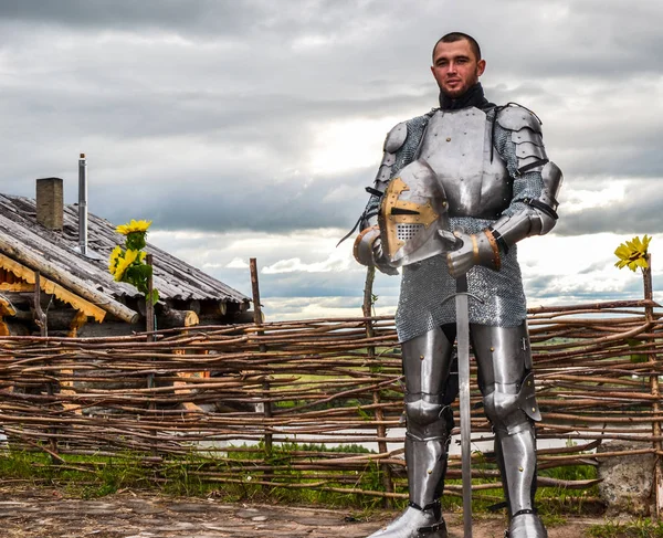 Knight in armor on the background of a wicker fence, a wooden house and the expanses of the motherland.