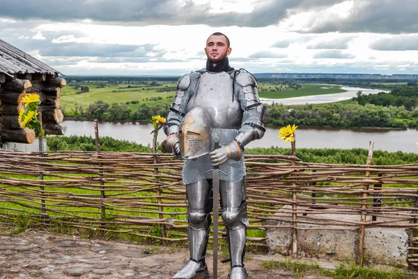 Knight in armor on the background of a wicker fence, a wooden house and the expanses of the motherland.