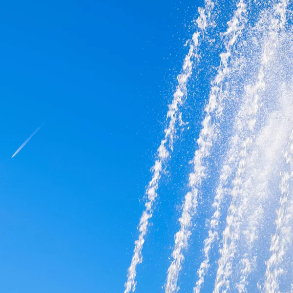 Jets and splashes of water fountain against the blue sky. Condensation track of a jet plane against the sky and a fountain.