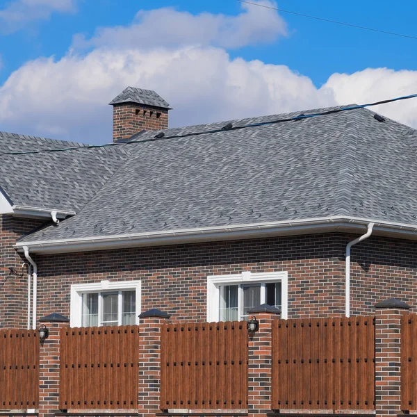 Torção de asfalto. Telhas decorativas de betume no telhado de uma casa de tijolos. Cerca feita de metal ondulado . — Fotografia de Stock