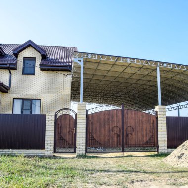 Bir çit ve gates ile tuğla ev. Çelik çerçeve ile büyük bir tente. Görünümü yeni bir İnşaatlı çit ve tuğla ve oluklu metal yapılmış bir ev.