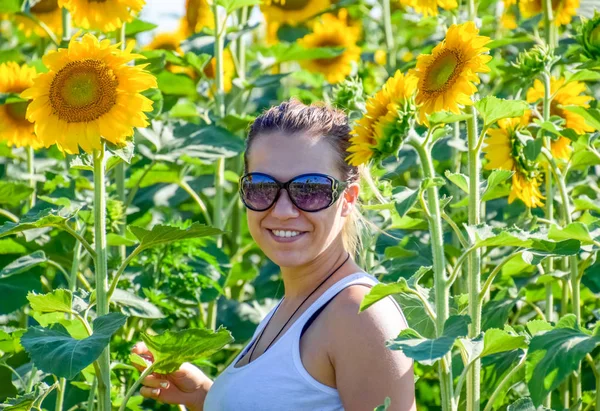Ξανθιά γυναίκα σε γυαλιά ηλίου και ένα λευκό t-shirt είναι χαμογελαστός ενώ στέκεται στο Ηλιοτρόπια. — Φωτογραφία Αρχείου