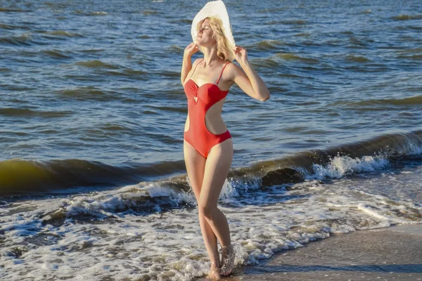 Κορίτσι με ένα λευκό καπέλο και σε ένα κόκκινο μαγιό στέκεται στην παραλία. — Φωτογραφία Αρχείου