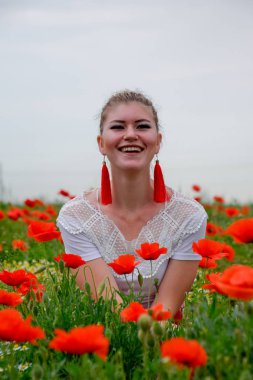 Blonde young woman in red skirt and white shirt, red earrings is in the middle of a poppy field. clipart