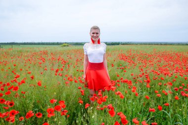 Kırmızı etekli, beyaz gömlekli, kırmızı küpeli sarışın bir kadın gelincik tarlasının ortasında..
