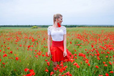 Kırmızı etekli, beyaz gömlekli, kırmızı küpeli sarışın bir kadın gelincik tarlasının ortasında..