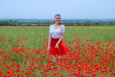 Kırmızı etekli, beyaz gömlekli, kırmızı küpeli sarışın bir kadın gelincik tarlasının ortasında..