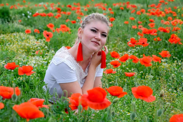 Jeune femme blonde en jupe rouge et chemise blanche, boucles d'oreilles rouges est au milieu d'un champ de pavot . — Photo