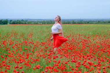 Kırmızı etekli, beyaz gömlekli, kırmızı küpeli sarışın bir kadın gelincik tarlasının ortasında..