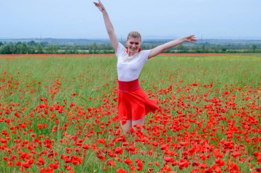 Kırmızı etekli, beyaz gömlekli, kırmızı küpeli sarışın bir kadın gelincik tarlasının ortasında..