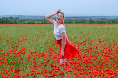 Kırmızı etekli, beyaz gömlekli, kırmızı küpeli sarışın bir kadın gelincik tarlasının ortasında..