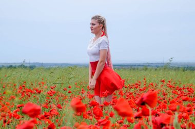 Kırmızı etekli, beyaz gömlekli, kırmızı küpeli sarışın bir kadın gelincik tarlasının ortasında..