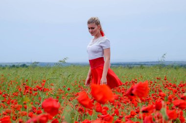 Kırmızı etekli, beyaz gömlekli, kırmızı küpeli sarışın bir kadın gelincik tarlasının ortasında..