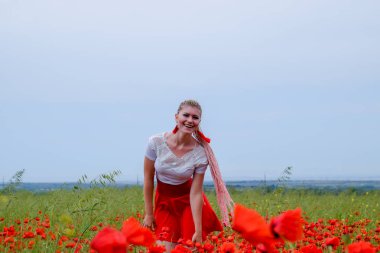 Kırmızı etekli, beyaz gömlekli, kırmızı küpeli sarışın bir kadın gelincik tarlasının ortasında..