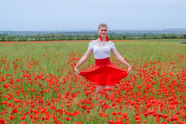 Kırmızı etekli, beyaz gömlekli, kırmızı küpeli sarışın bir kadın gelincik tarlasının ortasında..