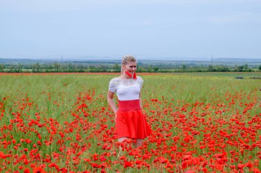 Kırmızı etekli, beyaz gömlekli, kırmızı küpeli sarışın bir kadın gelincik tarlasının ortasında..