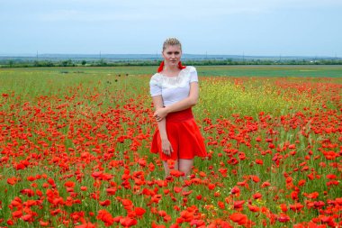 Kırmızı etekli, beyaz gömlekli, kırmızı küpeli sarışın bir kadın gelincik tarlasının ortasında..