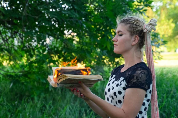 girl holds a burning book in her hands. A young woman in a forest burns a book.