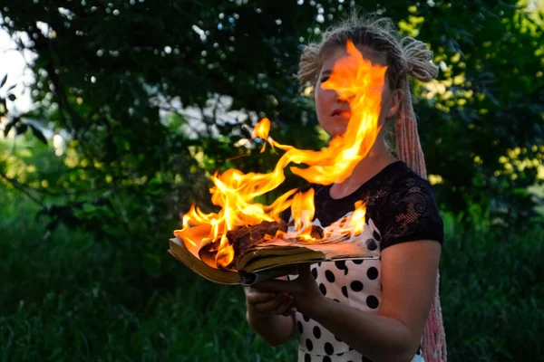 Girl holds a burning book in her hands. A young woman in a forest burns a book. — Stock Photo, Image