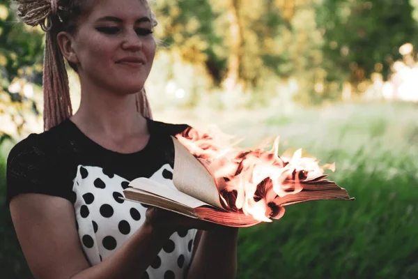 Fille tient un livre brûlant dans ses mains. Une jeune femme dans une forêt brûle un livre . — Photo