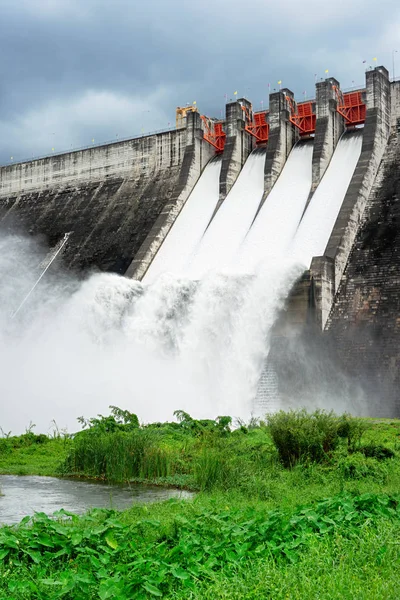 Concrete dam with flowing water from spillways with green grass and cloudy sky.