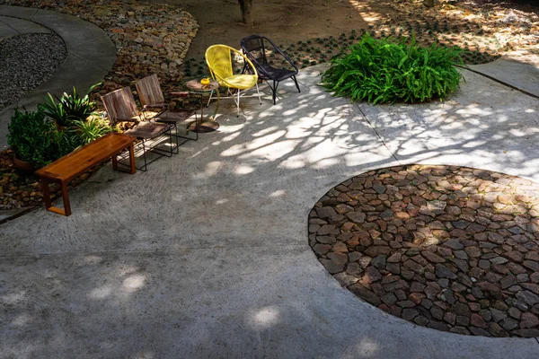 Wooden chairs and bench in garden with stones and concrete floor and shadow of the trees.