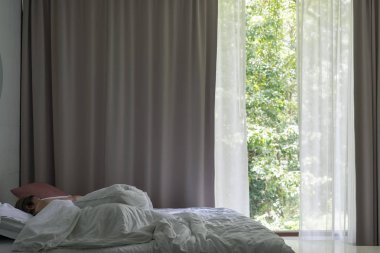 Asyalı kadın büyük pencere perdeleri ve yeşil orman arka plan ile yatakta uyuyor.