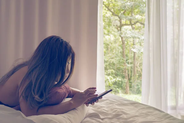 Asyalı kadın pencere perde ve ağaçlar arka plan ile hareket eden telefon içinde elleri bakarak yatakta yalan.