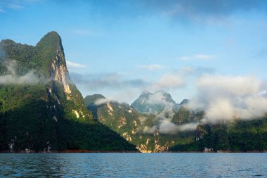 Sabah ışığında yeşil kireçtaşı sıradağları sis, bulutlar, göl ve mavi gökyüzü Tayland, Surat Thani 'deki Khao Sok Ulusal Parkı' nda..