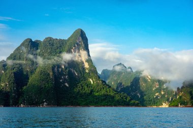 Tayland, Surat Thani 'deki Khao Sok Ulusal Parkı' nda mavi göllü yeşil kireçtaşı sıradağları ve ılık sabah ışığında beyaz bulutlar..