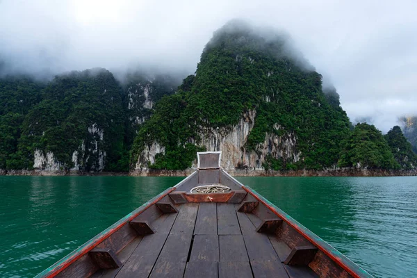 Wooden boat in the lake with limestone mountain in the morning mist and clouds in Surat Thani, Thailand.