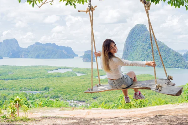 Asian woman smiling and sitting on a wooden swing with limestone islands and sea background in Phang Nga, Thailand.