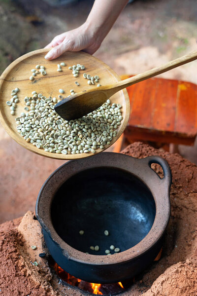 Hand holding wooden tray with raw coffee bean and clay pot on fire stove.