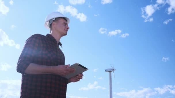 Der männliche Ingenieur inspiziert das Windrad auf dem Tablet. Sonniger Tag und Wolken. Ein Mann trägt ein kariertes Hemd und einen weißen Bauhelm. — Stockvideo