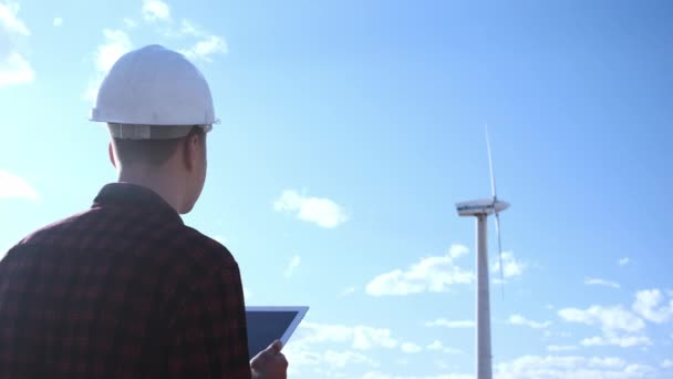 Der männliche Ingenieur inspiziert das Windrad auf dem Tablet. Sonniger Tag und Wolken. Ein Mann trägt ein kariertes Hemd und einen weißen Bauhelm. — Stockvideo