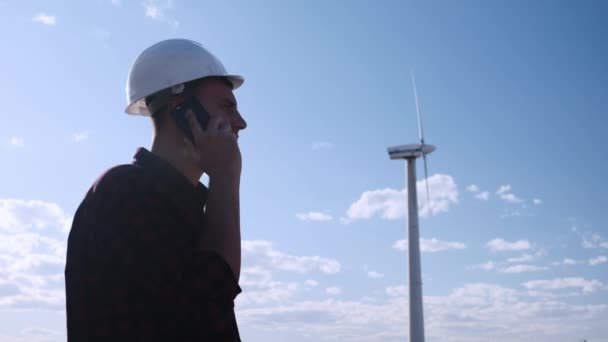 Инженер-мужчина осматривает ветряную турбину на планшете. Солнечный день и облака. Мужчина одет в клетчатую рубашку и в белый строительный шлем . — стоковое видео