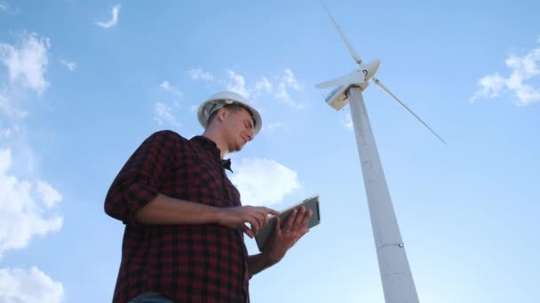 Der männliche Ingenieur inspiziert das Windrad auf dem Tablet. Sonniger Tag und Wolken. Ein Mann trägt ein kariertes Hemd und einen weißen Bauhelm. — Stockvideo