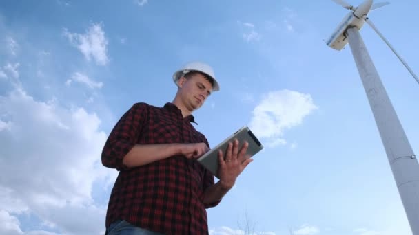 남성 엔지니어는 태블릿에 바람 터빈을 검사 합니다. 화창한 날 그리고 구름입니다. 남자는 옷을 입고 격자 무늬 셔츠와 엔지니어 흰색 건설 헬멧. — 비디오