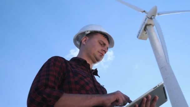 Mužské inženýr zjišťuje Větrná turbína na tabletu. Slunečný den a mraky. Muž je oblečená v kostkované košili a bílé stavbařskou přílbu inženýři. — Stock video