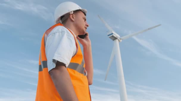 L'ingegnere maschio proietta il lavoro vicino alla turbina eolica. Fa una telefonata. Giorno soleggiato e nuvole. L'uomo è vestito con un giubbotto rosso e un casco bianco da ingegnere. . — Video Stock