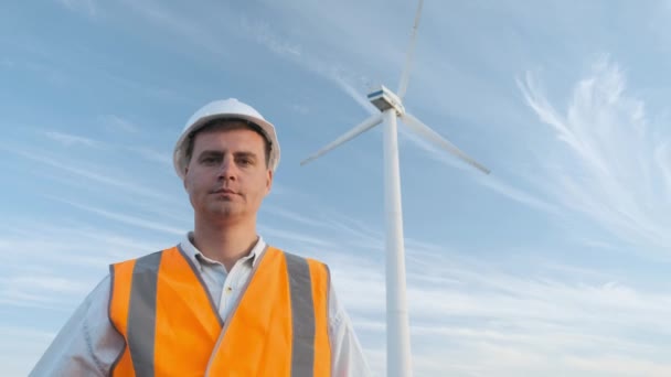 Portrét: mužské inženýr projektů práci poblíž větrné turbíny. Slunečný den a mraky. Ten člověk je oblečený v červené brýle vestu a helmu inženýři bílé. — Stock video
