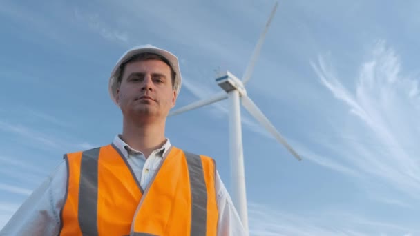 Retrato: o engenheiro masculino projeta o trabalho próximo à turbina eólica. Dia ensolarado e nuvens. O homem está vestido com um colete de vidro vermelho e um capacete branco de engenheiros . — Vídeo de Stock