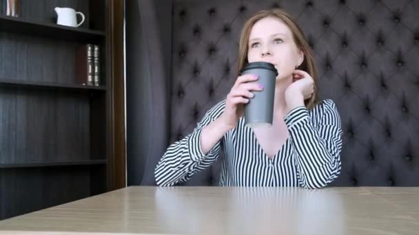 Ein junges schönes Mädchen in einem stilvollen Café trinkt morgens vor der Arbeit Kaffee. sitzt dem Fenster gegenüber. Er trinkt Kaffee aus einem schwarzen Pappglas. Nahaufnahme. — Stockvideo