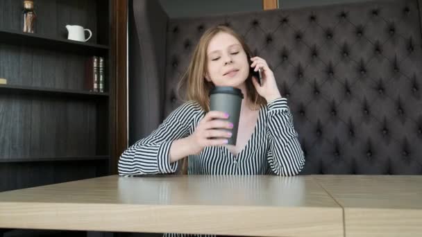 Ung vacker flicka kallar vid smartphone i caféet. Hon dricker morgon kaffe innan arbetet. Koncept: Frukost, koppla av, kaffe, business — Stockvideo