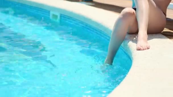 Menina feliz em um maiô preto e um chapéu branco se senta na borda de uma piscina ao ar livre. Mete o pé na água. Um verdadeiro descanso à beira da piscina. Recurso no mar. Descanso rico — Vídeo de Stock