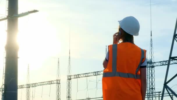 En flicka ingenjör i en vit hjälm och en orange skyddande väst inspekterar ett kraftverk med sladdar och elektricitet. Löser frågan om nya ägare. — Stockvideo