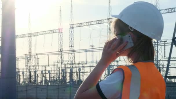 En flicka ingenjör i en vit hjälm och en orange skyddande väst inspekterar ett kraftverk med sladdar och elektricitet. Löser frågan om nya ägare. — Stockvideo