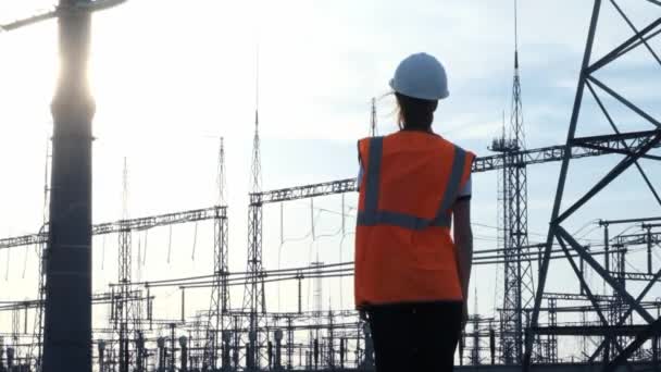 Una niña ingeniera con un casco blanco y un chaleco protector naranja inspecciona una central eléctrica con cables y electricidad. Resuelve la cuestión de la nueva propiedad . — Vídeos de Stock